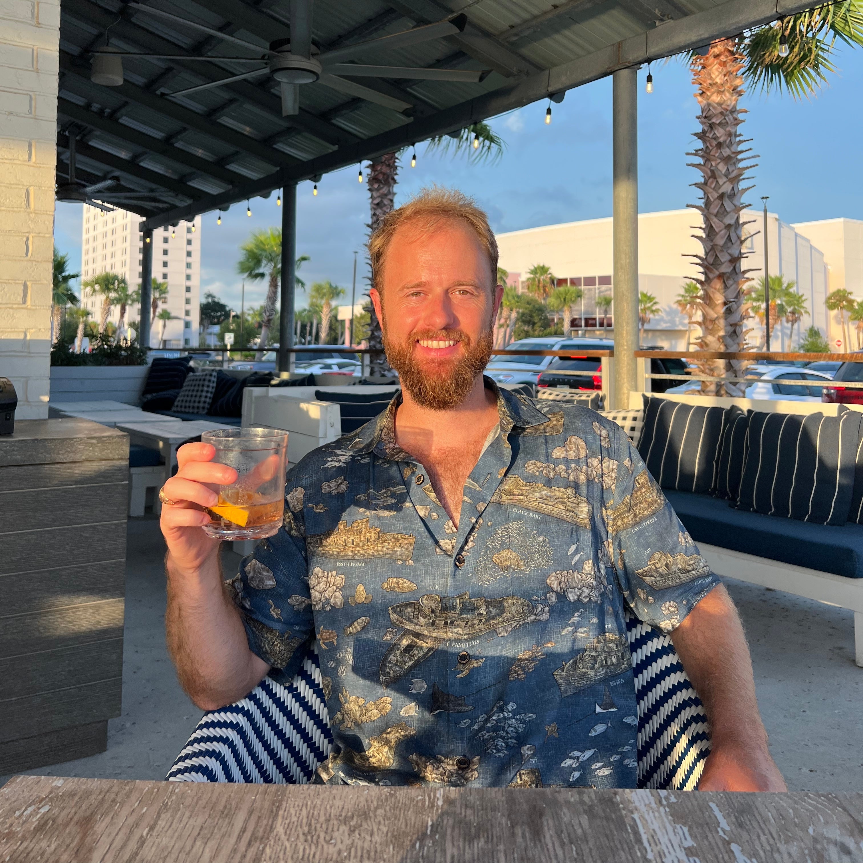 A man wearing the Dive PCB Floridian Shirt by Okihasi sitting at a table holding a drink.